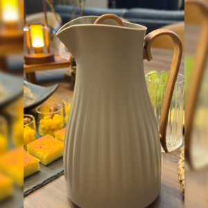 Garrafa térmica para café na cor café sobre uma mesa posta arrumada com uma bandeja de bolos, uma lanterna e um copo de vidro.