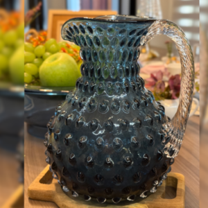 Jarra Estilo Murano na cor azul com detalhes em relevo e alça translúcida em uma mesa posta, com frutas ao fundo