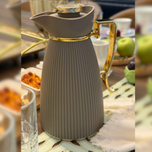 Garrafa Térmica Zaya em tonalidade café com detalhes dourados em sua alça e ampola de vidro em uma mesa posta.